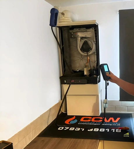 An open wall-hung boiler in a domestic kitchen with its front case off: the fan and heat exchanger exposed, a gas valve and pipework below them and a red readout on the control strip. A hand and forearm at frame right hold a handheld analyser with a lit screen; no head and no face is in frame. On the floor beneath, the firm's own branded mat carries its mark, its website address, its mobile number and its six-digit registration number. A screwdriver lies on the mat. A tiled splashback, a wood floor and a grey blind at the right edge.