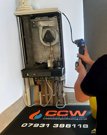 An open wall-hung boiler with its front case off: the fan and heat exchanger exposed, a digital readout on the control strip, and the pipe tails and isolating valves below it. A person in a dark T-shirt stands at frame right holding a handheld analyser in both hands, its probe lead running up into the boiler; no head and no face is in frame. The firm's own branded mat lies beneath, carrying its mark, its website address, its mobile number and its six-digit registration number. A yellow tool case sits in the bottom right corner. A tiled skirting and a plain wall behind.